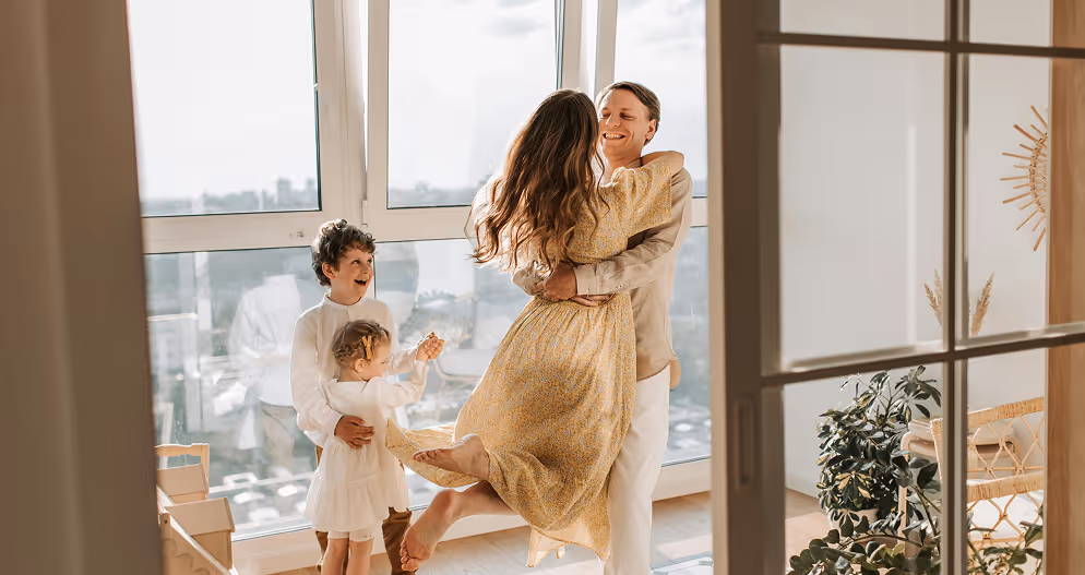 A joyful family in a bright room with large windows; a man lifts a woman in a yellow dress while two children watch and smile.