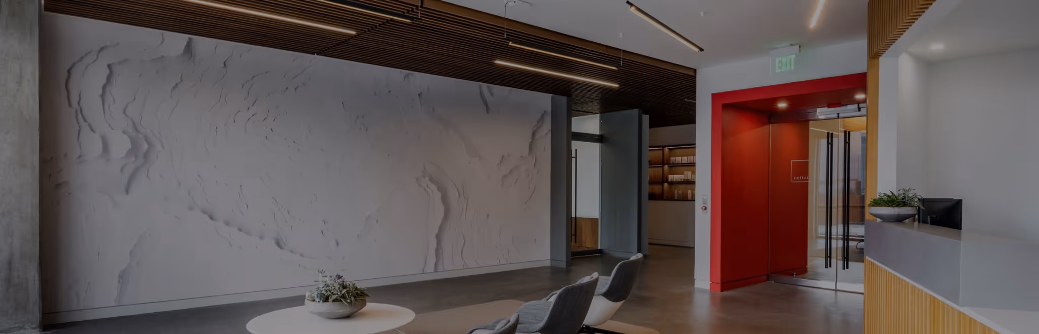 Modern office lobby with textured white feature wall, wood slat ceiling, linear lighting, and a red glass meeting room entrance.
