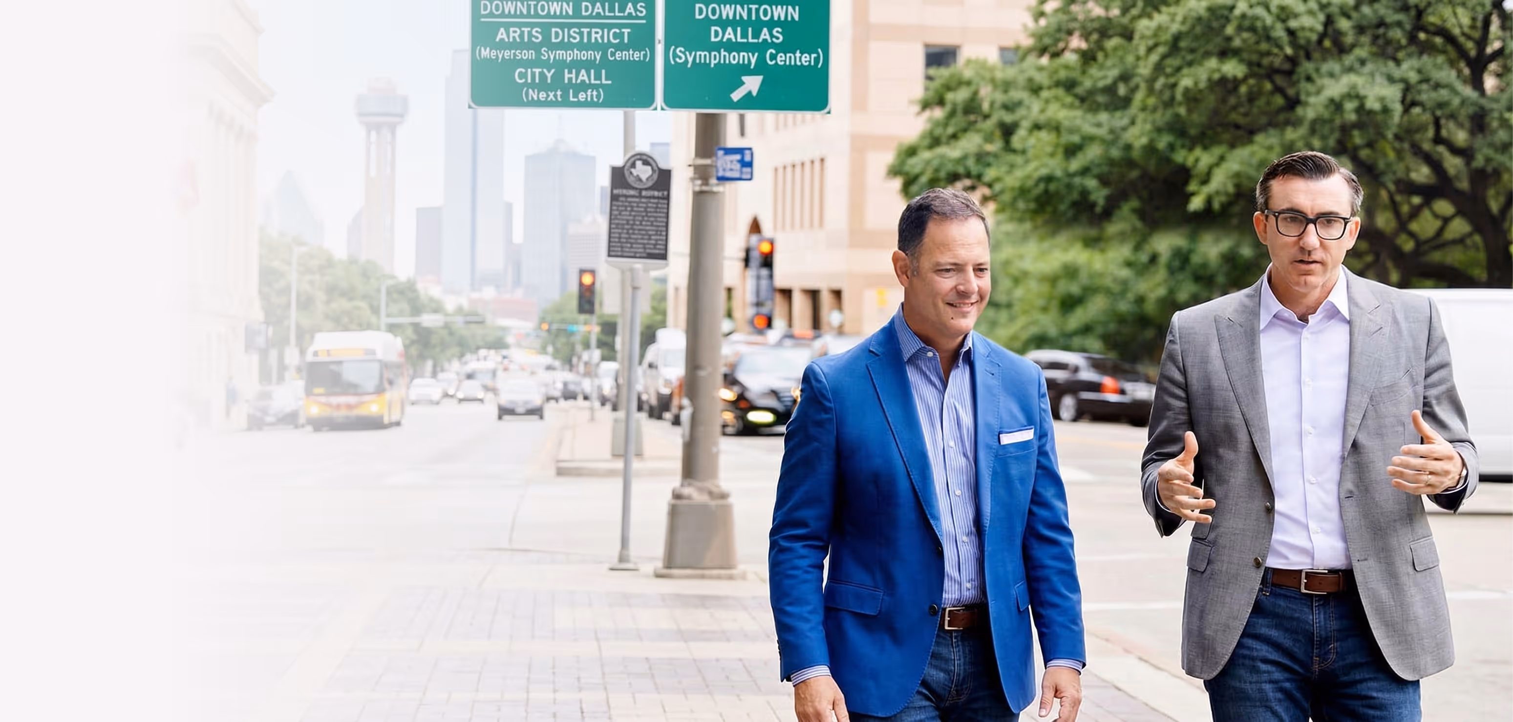 Two men in business casual attire walking and talking on a downtown Dallas sidewalk under street signs.