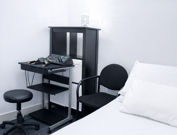 Clinical room with a black examination chair, a medical device on a small metal cart, and a white bed with two pillows.