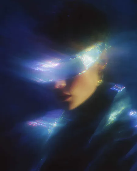 Portrait of a person wearing a glowing, futuristic visor and jacket in a dark, blue-lit environment.