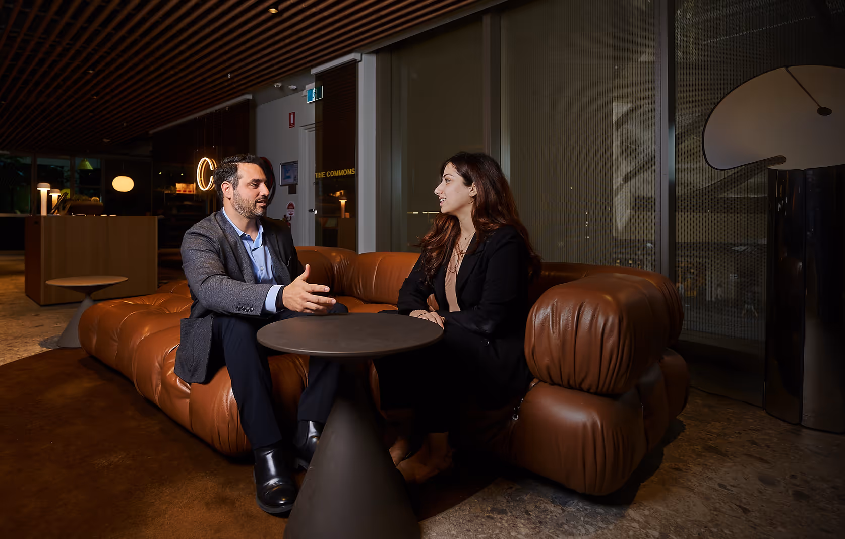 Man and woman in business attire sitting on a brown leather couch, having a conversation across a small round table in a modern office lounge.