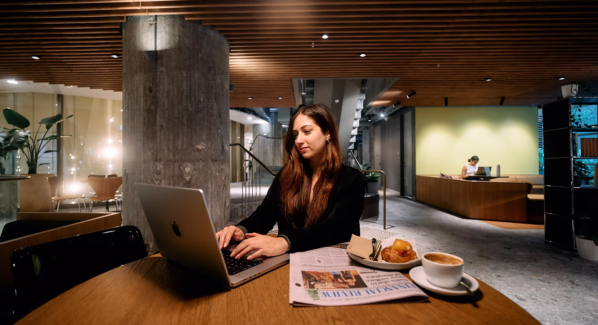 Young woman working on a laptop at a wooden table with a newspaper, pastry, and coffee in a modern cozy cafe.