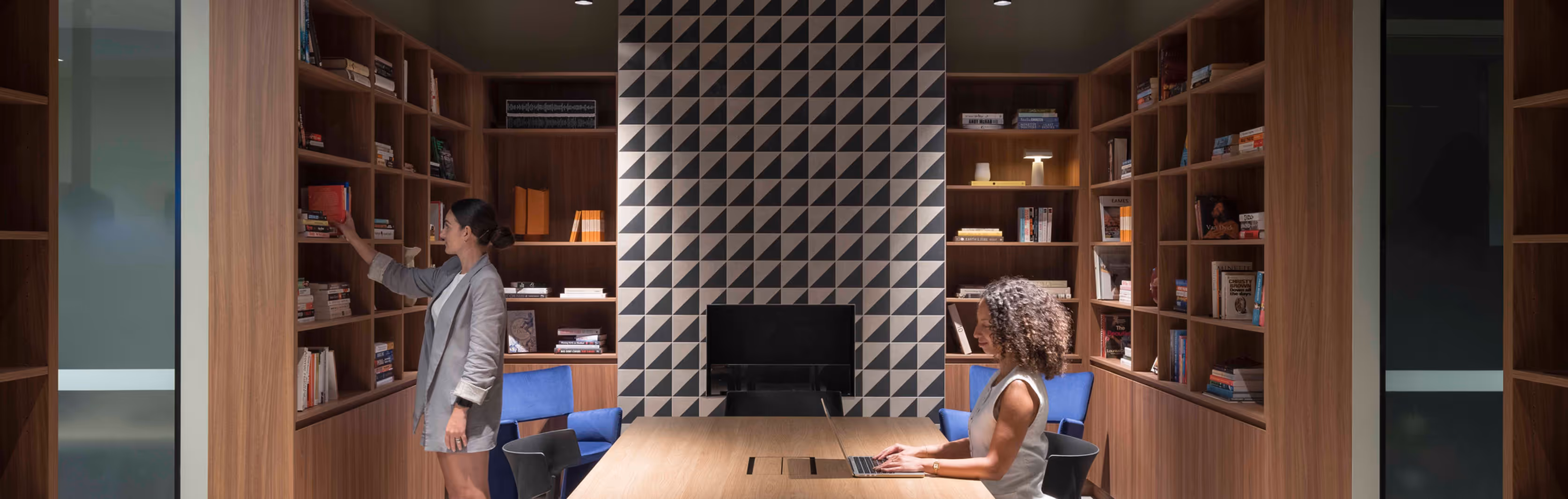 Two women in a modern office library; one woman is selecting a book from a wooden bookshelf, while the other is sitting at a wooden table working on a laptop.