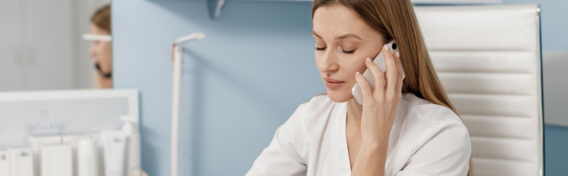 Woman in white coat sitting in an office chair, speaking on a smartphone with eyes closed.