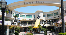 Outdoor shopping plaza with stores, palm trees, lampposts, and a yellow umbrella over an outdoor seating area under a curved sign reading "Encino Place."