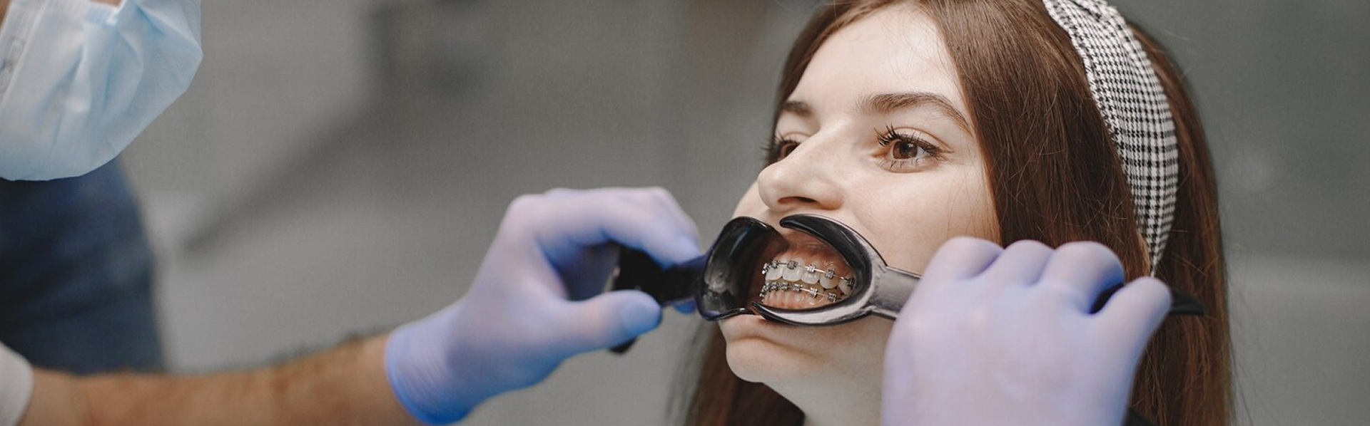 Dentist with gloves placing a cheek retractor in a young woman's mouth showing her braces.
