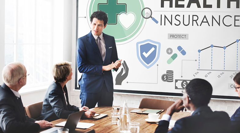 Businessman in a blue suit presenting health insurance concepts on a screen to a group of four professionals around a conference table.