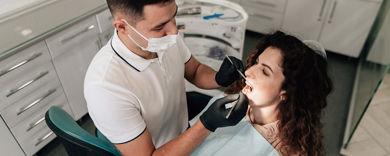 Dentist wearing a mask and gloves examining the teeth of a woman reclining in a dental chair.