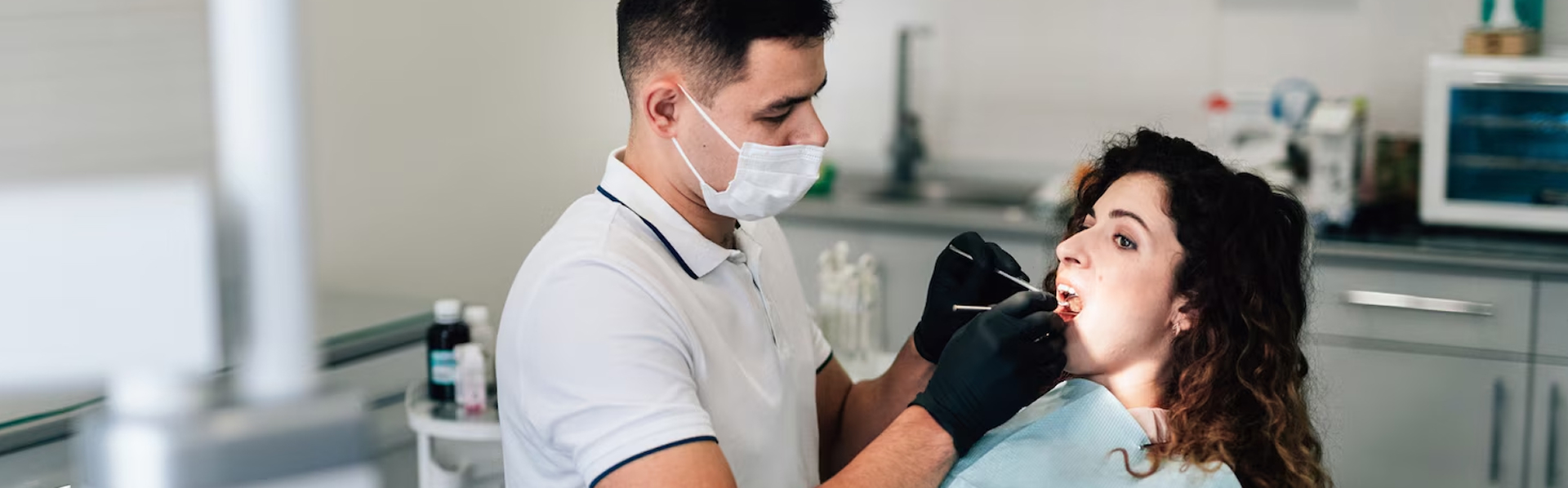 Dentist wearing a mask and gloves examining a female patient's mouth in a dental clinic.
