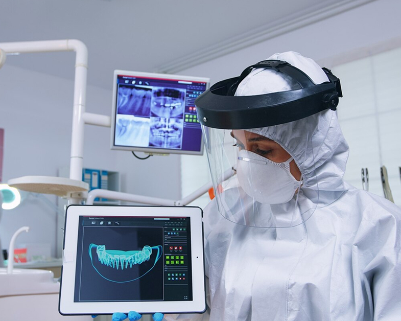 Dental professional wearing full protective gear and face shield, holding a tablet displaying a 3D digital model of teeth in a dental clinic.
