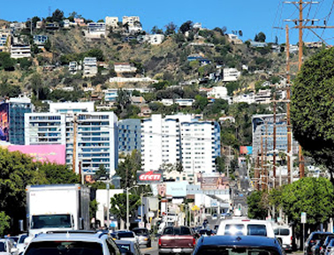 Busy city street with cars, buildings, and hills covered with houses in the background under a clear blue sky.