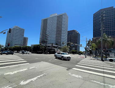 City intersection with traffic lights, crosswalks, cars, and tall office buildings under a clear blue sky.