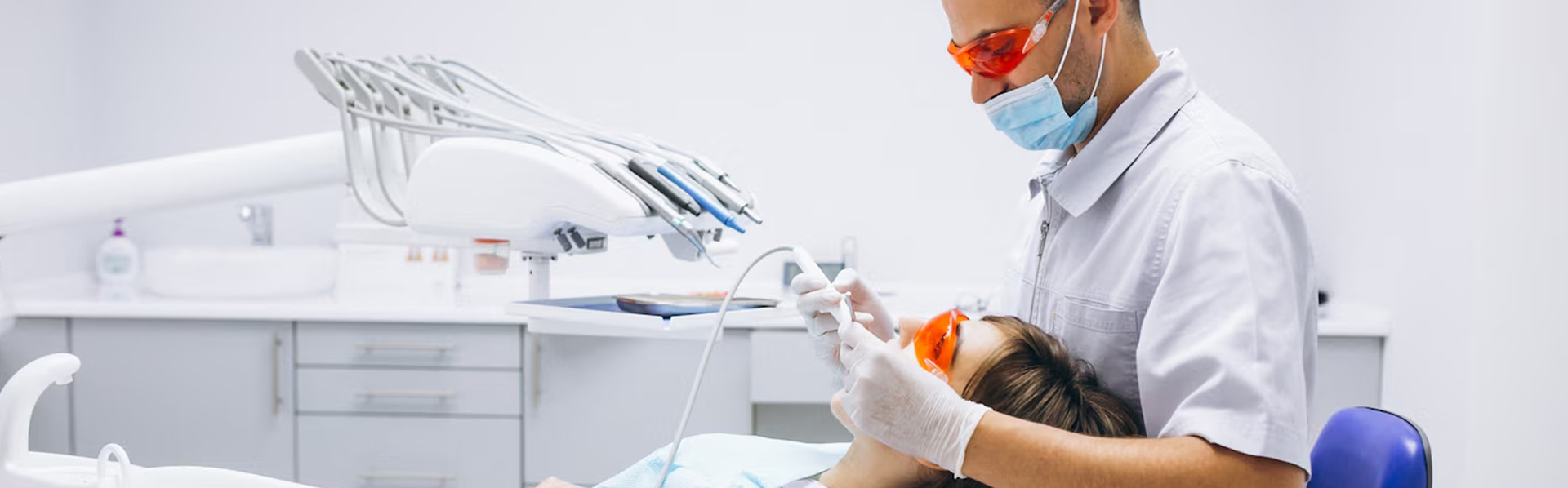 Dentist wearing protective glasses and mask treats patient lying in dental chair with orange protective glasses in a modern clinic.