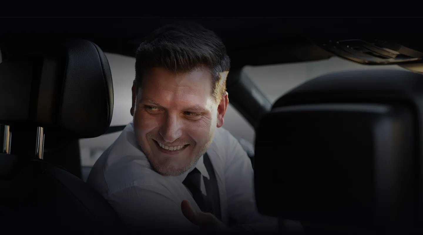 Smiling man in white shirt and black tie seated inside a car, looking back over his shoulder.