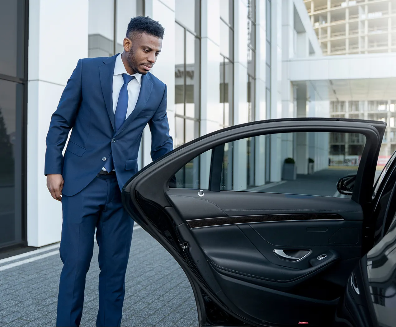Man in a blue suit holding open the rear door of a black car outside a modern office building.
