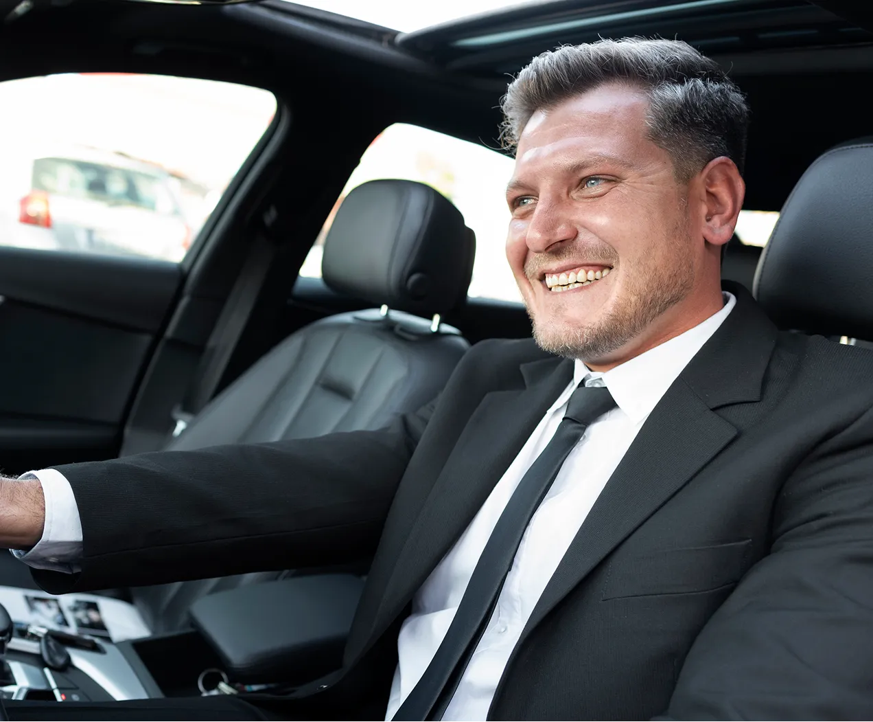 Smiling man in a black suit driving a car with leather seats.