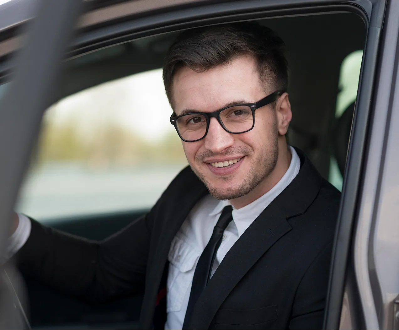 Smiling man with glasses wearing a suit and tie sitting in the driver's seat of a car.