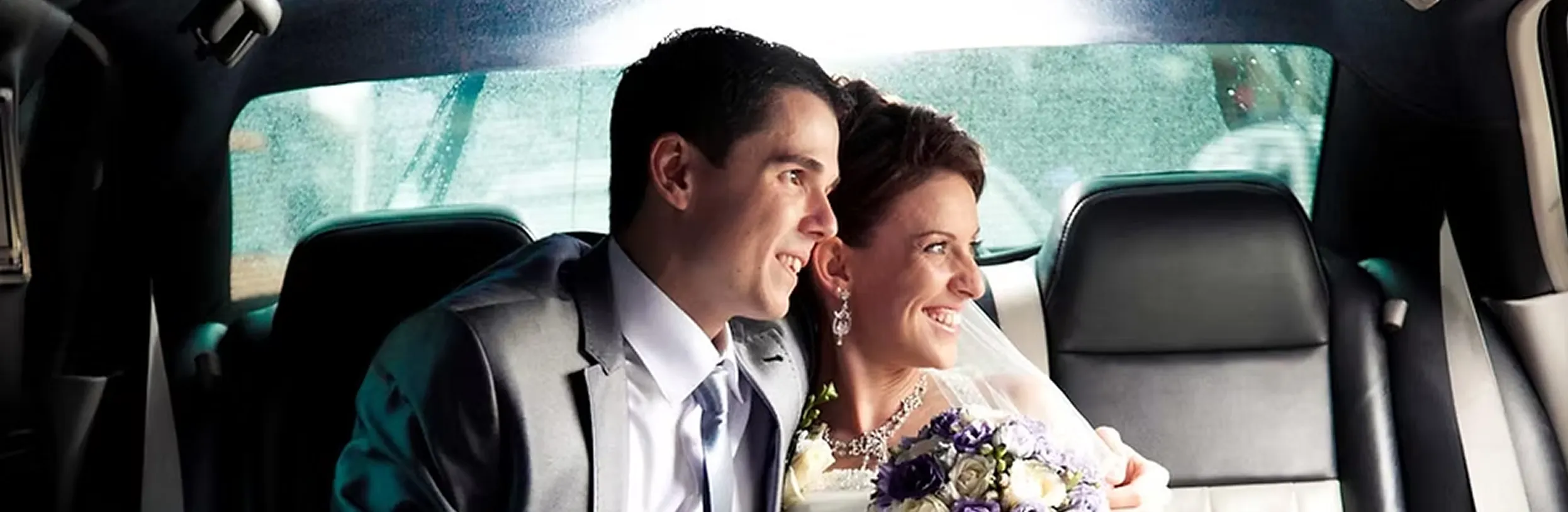 Smiling bride and groom sitting closely in the backseat of a car, bride holding a bouquet of white and purple flowers.