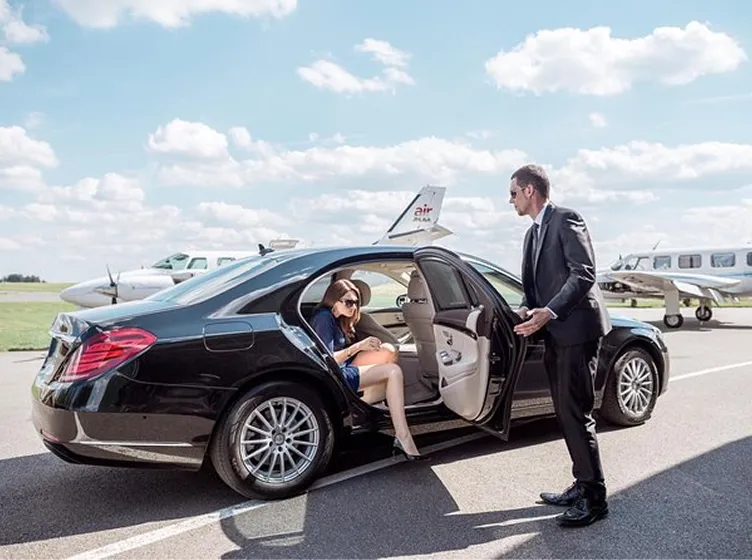 Woman wearing sunglasses stepping out of a black luxury car with the help of a man in a suit at an airport with small airplanes in the background.