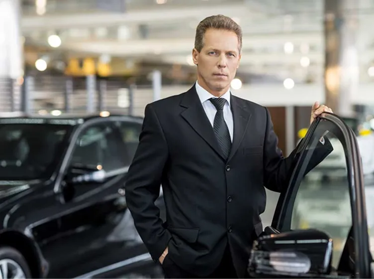 Man in black suit standing beside an open car door in a modern parking garage.