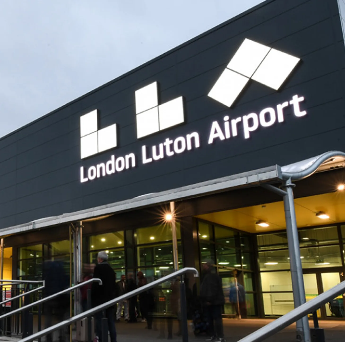Entrance of London Luton Airport with illuminated logo and people walking near the glass doors.