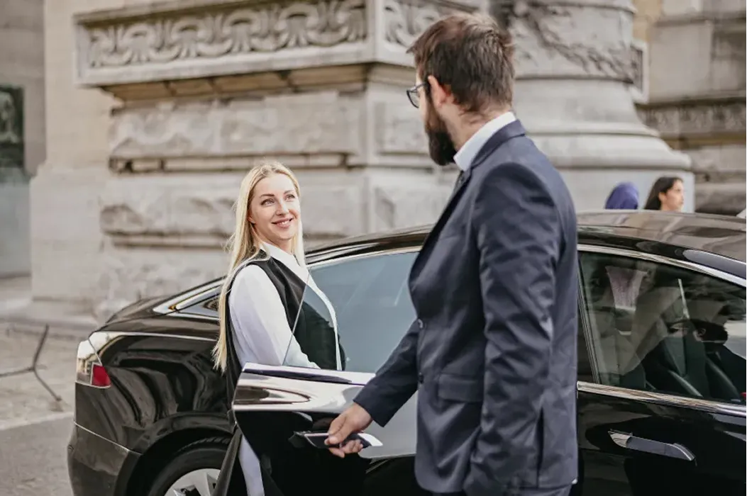 Man in suit opening car door for smiling woman standing beside a black luxury car in an urban setting.