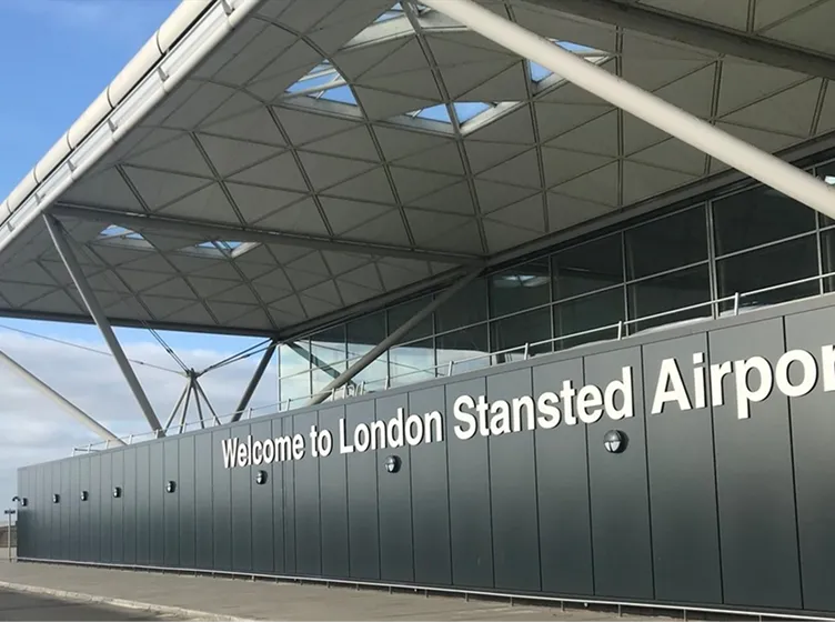 Exterior view of London Stansted Airport with large sign reading 'Welcome to London Stansted Airport' on gray panels under a modern canopy roof.