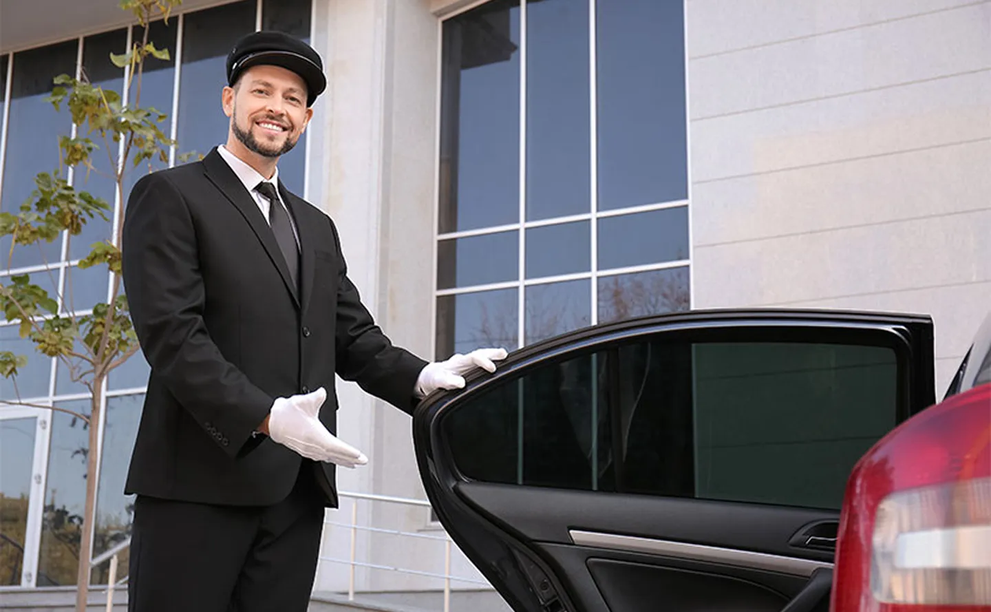 Chauffeur in black suit and white gloves holding open a car door outside a building.