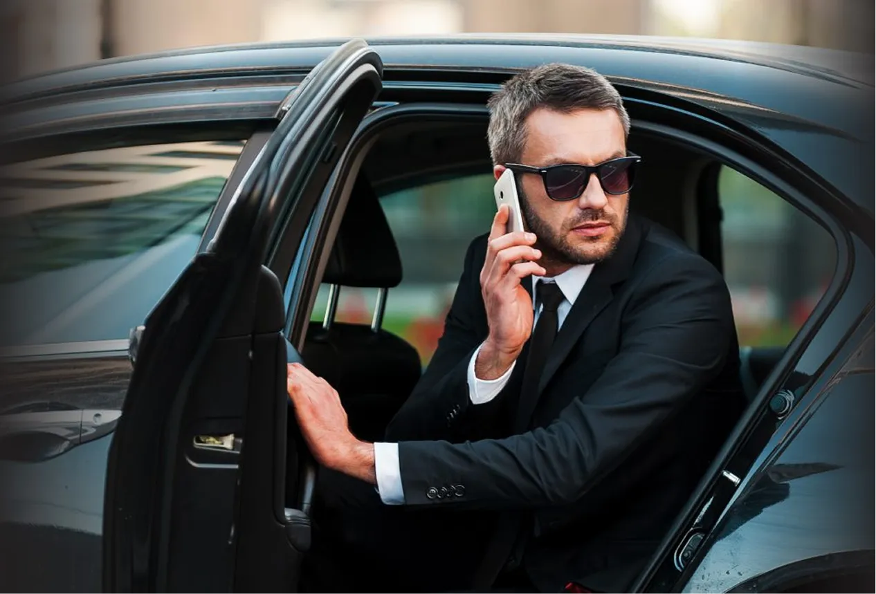 Man in a black suit and sunglasses sitting in a black car's back seat while talking on a smartphone.