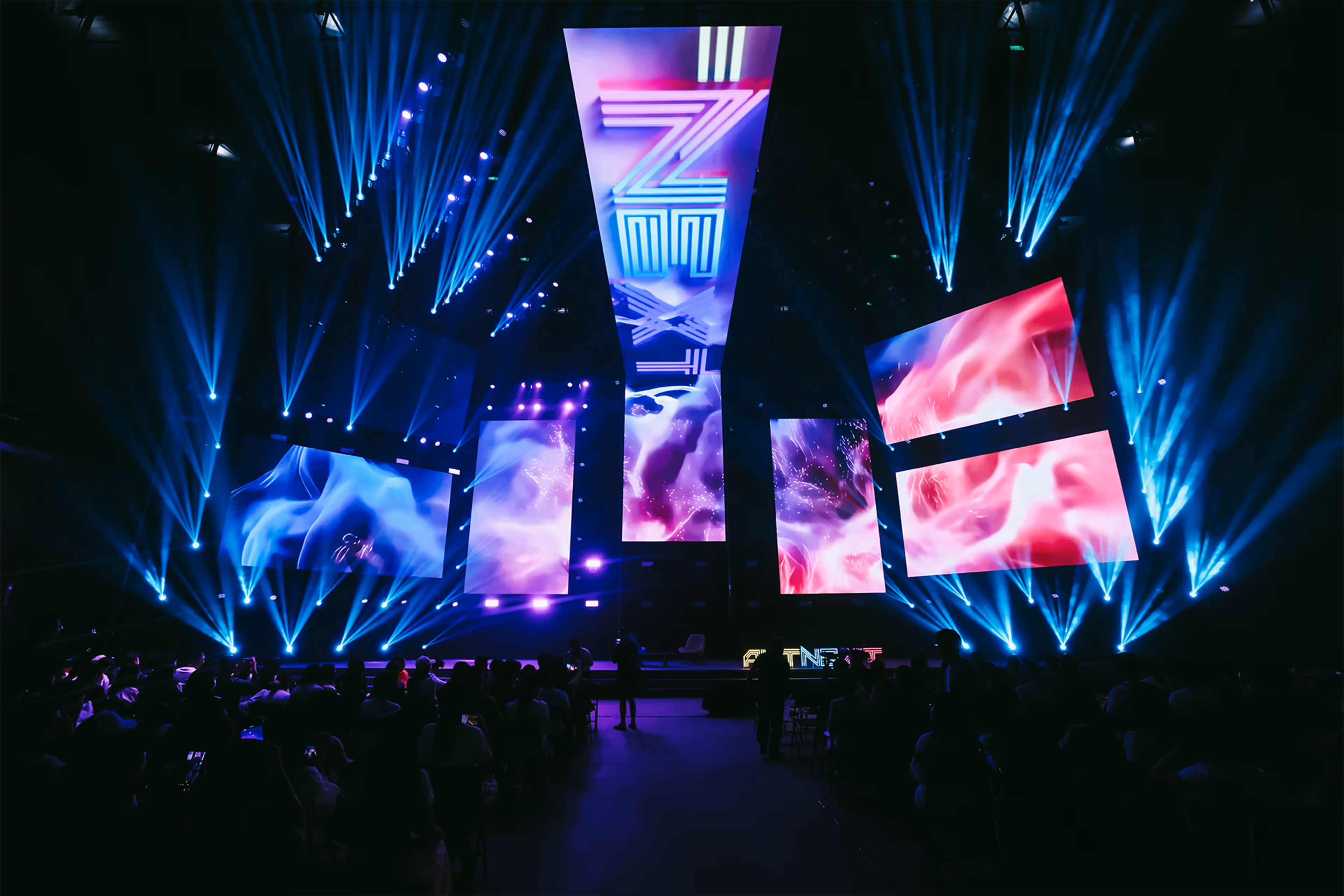 Dark event stage illuminated by blue spotlights with six large vertical screens displaying abstract purple and pink visuals and a crowd seated in front. This was the setup of AltNext Season 1.
