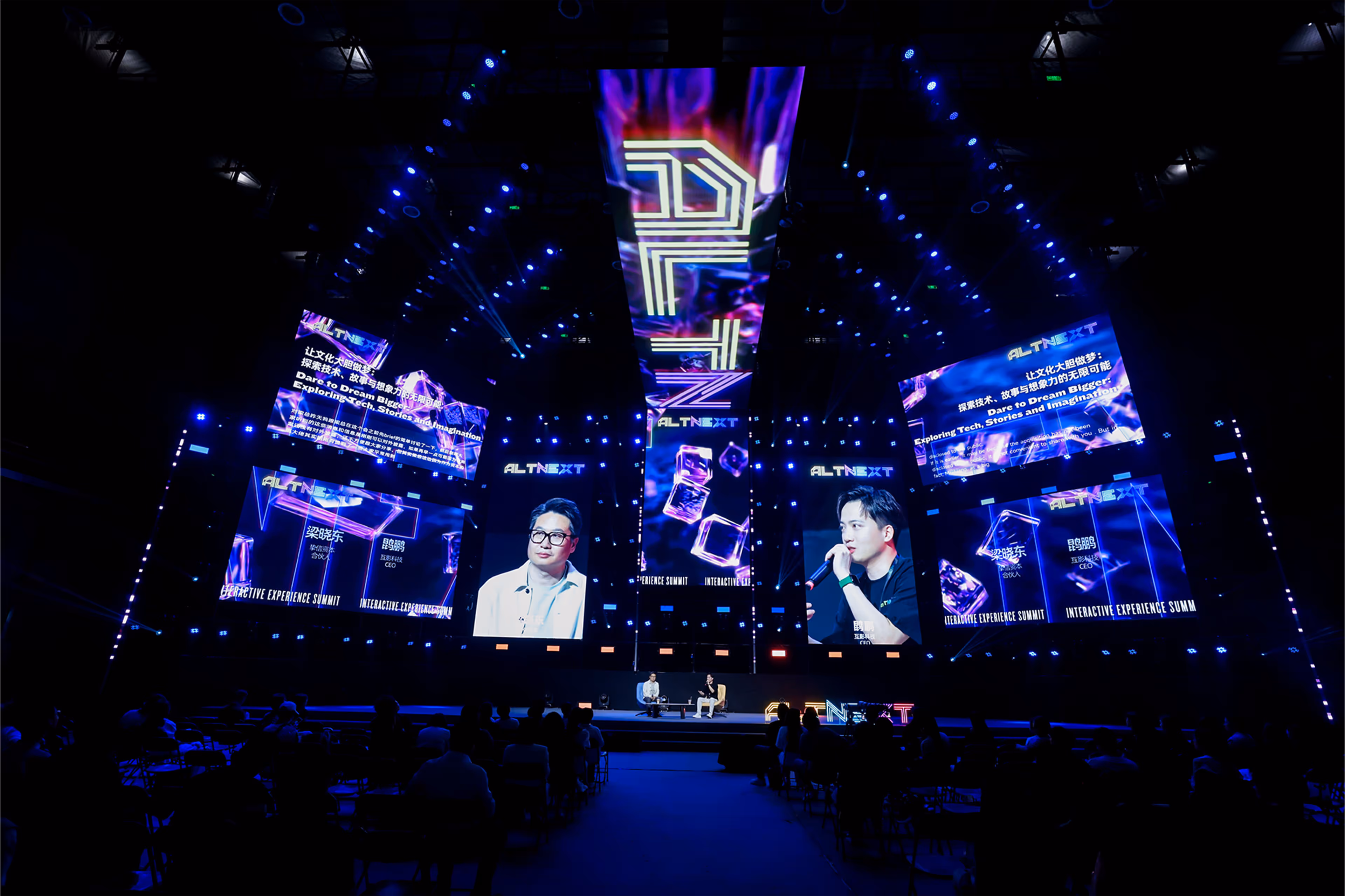 Two men seated on a stage having a discussion at the ALTNEXT Interactive Experience Summit with large digital screens and blue lighting in an auditorium. One of the men is Gary Kun, CEO of AltNext.