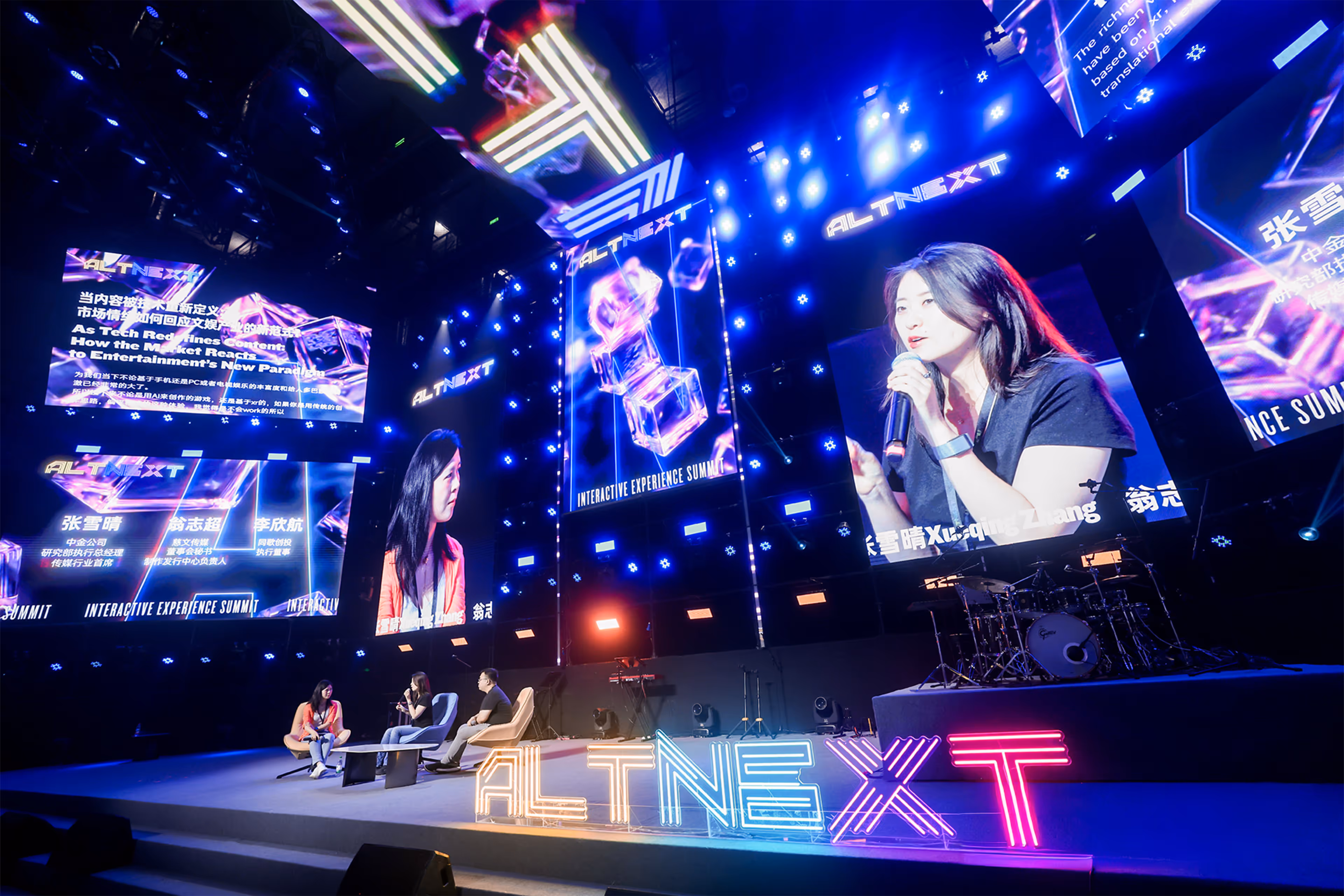Three people sitting on stage chairs engaged in a panel discussion at the ALTNEXT Interactive Experience Summit with large digital screens behind them.