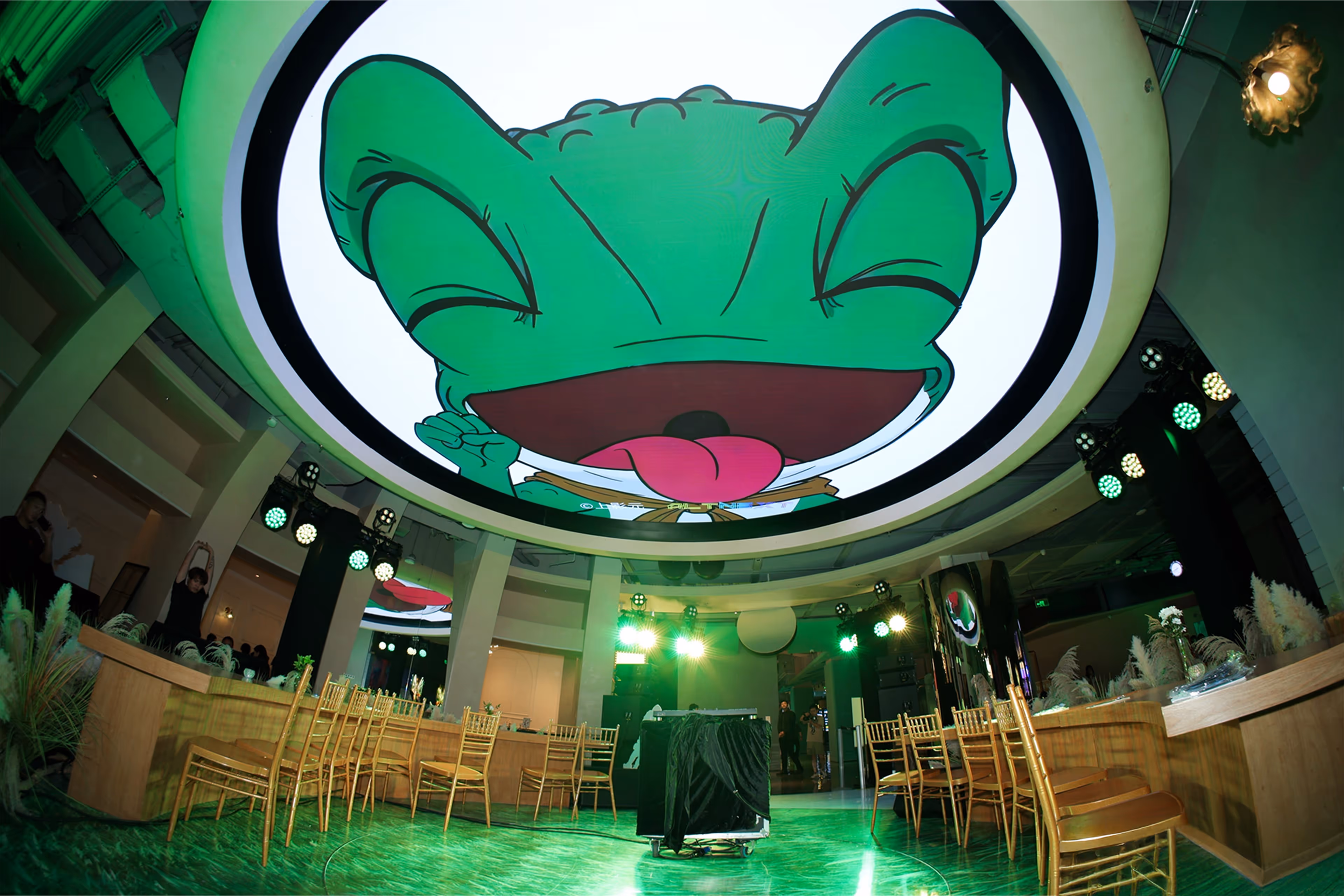 Interior of an event space with a large circular ceiling screen displaying a green cartoon frog with its tongue out - at AltNext Edition 1 in 2025.