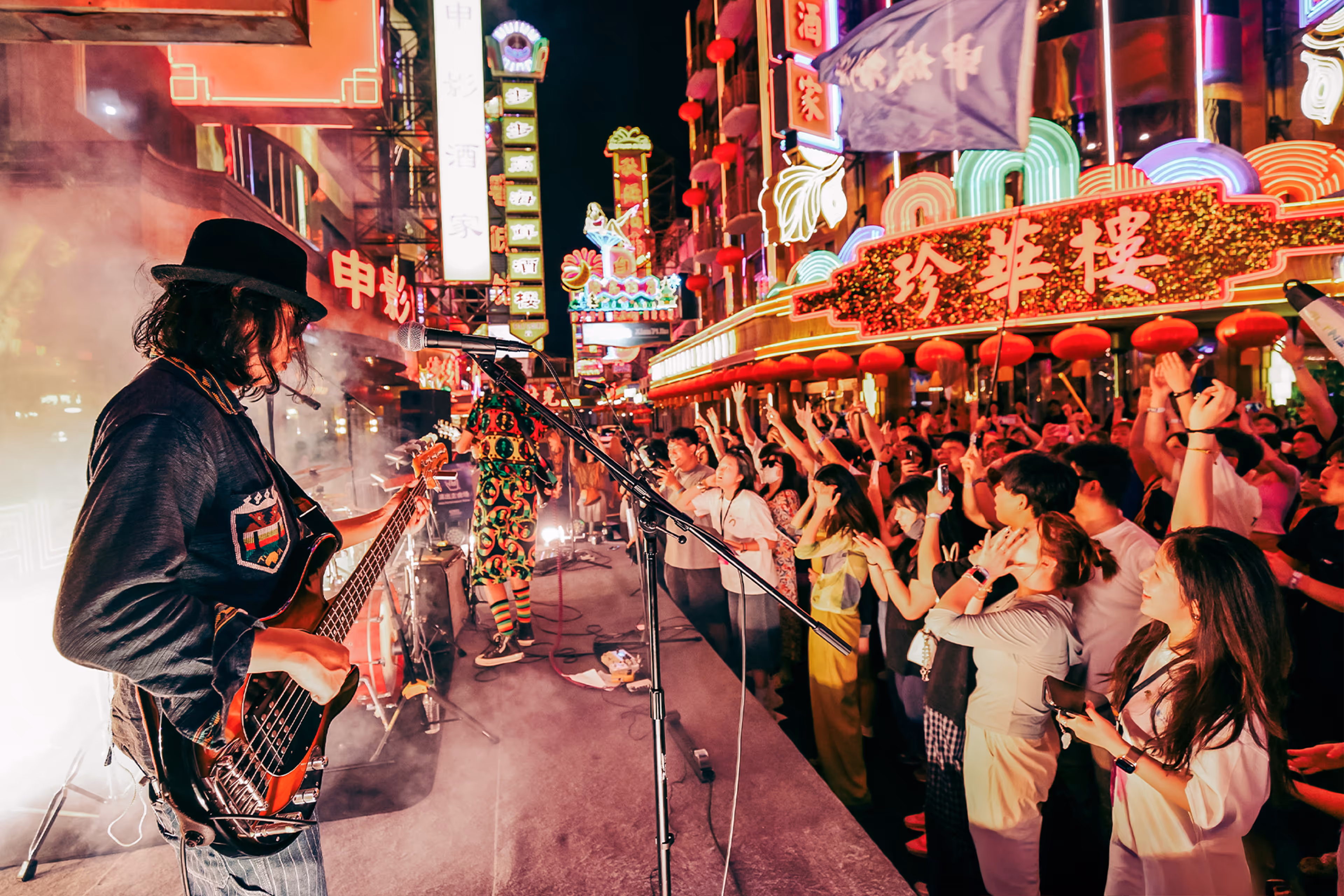 Musician playing electric bass guitar on stage at a lively night concert with colorful neon signs and an enthusiastic crowd raising hands - at AltNext Edition 1 in 2025.