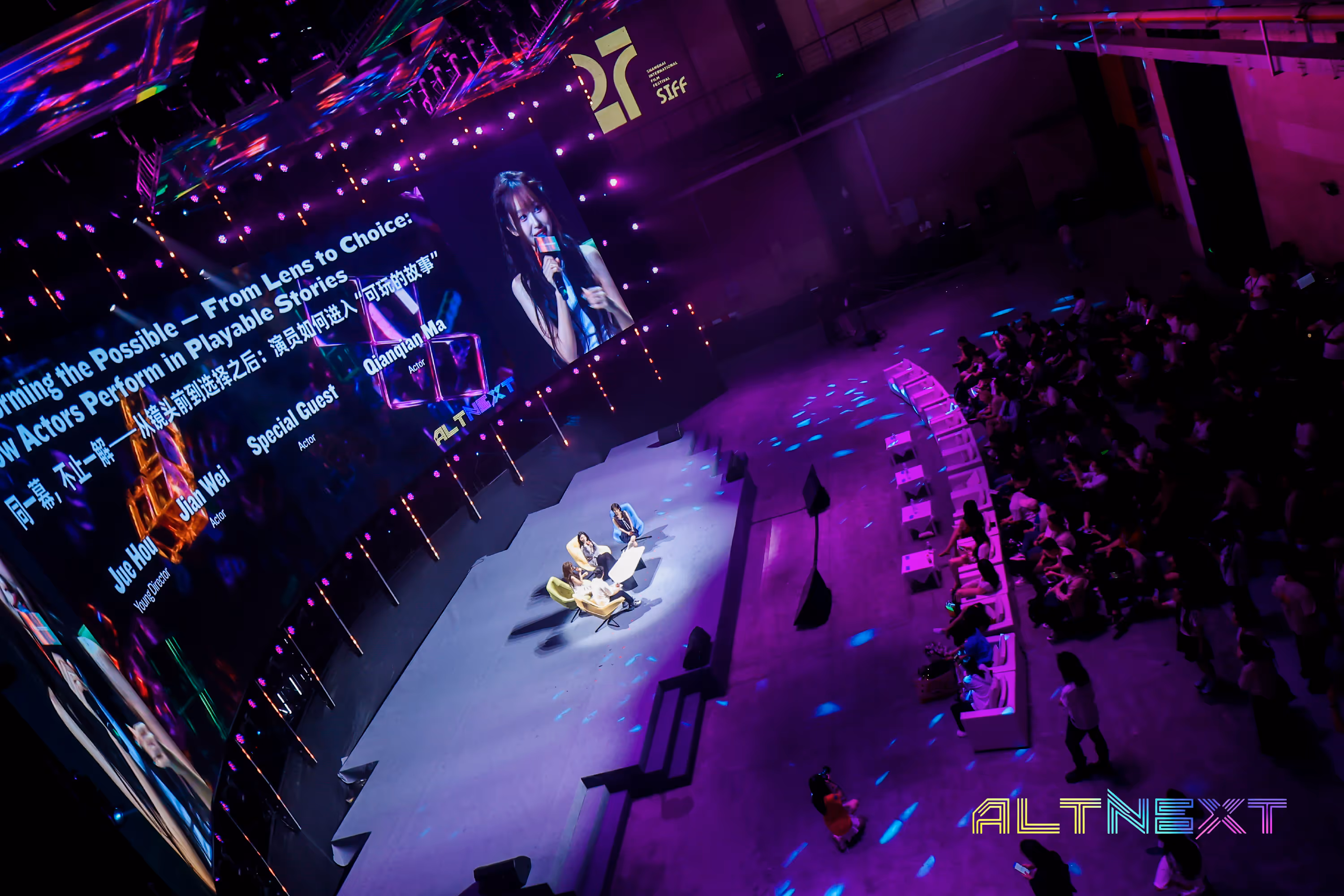 Panel discussion at a film festival with three people seated on stage under bright lights, a large screen showing a female speaker, and an audience seated in front under purple lighting - at AltNext Edition 1 in 2025.
