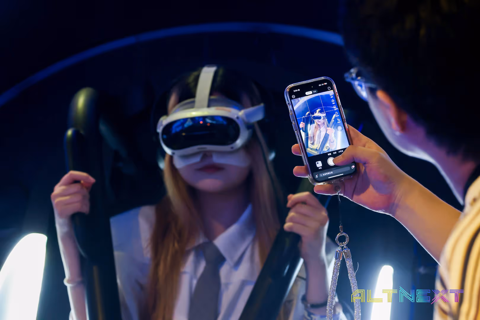 A person wearing a virtual reality headset and holding handles is being photographed or recorded on a smartphone by another person in a dark environment with neon lighting - at AltNext Season 1 (2025).
