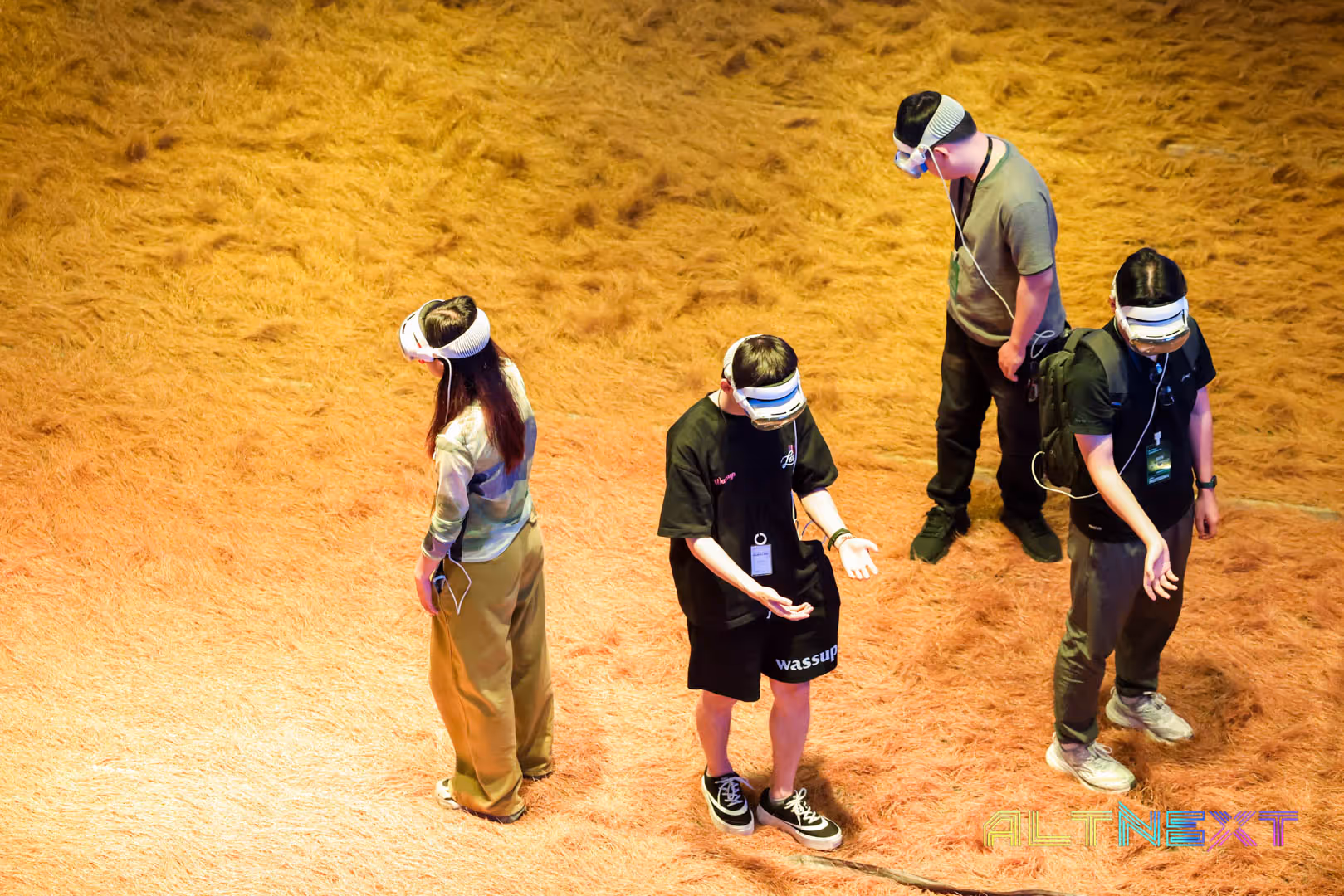 Four people wearing VR headsets and earphones standing on a textured orange floor under warm lighting - at AltNext Season 1 (2025).