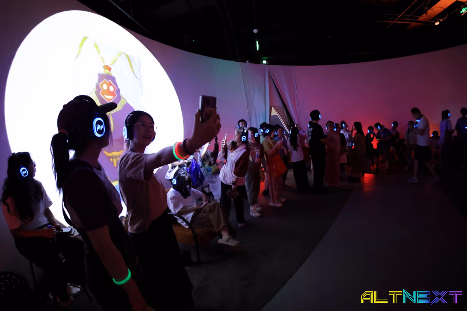 Crowd wearing glowing headphones at a dark indoor event with a colorful animated projection on the wall - at AltNext Season 1 (2025).