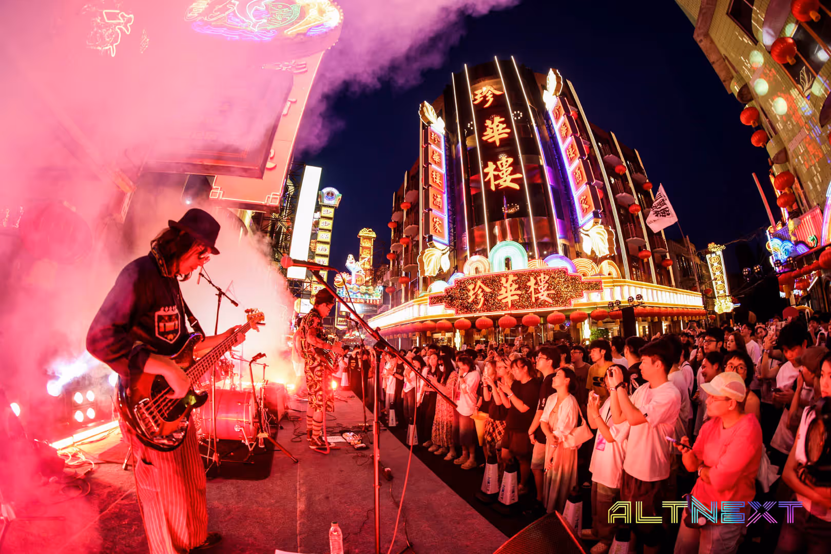Live band performing on an outdoor stage at night with colorful neon-lit buildings and a crowd watching - at AltNext Season 1 (2025).