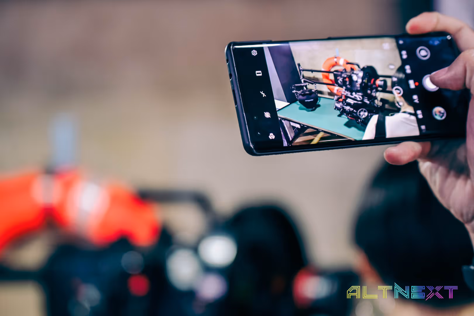 Hand holding a smartphone capturing a photo of a camera setup on a green table with an interactive display in the background - at AltNext Season 1 (2025).