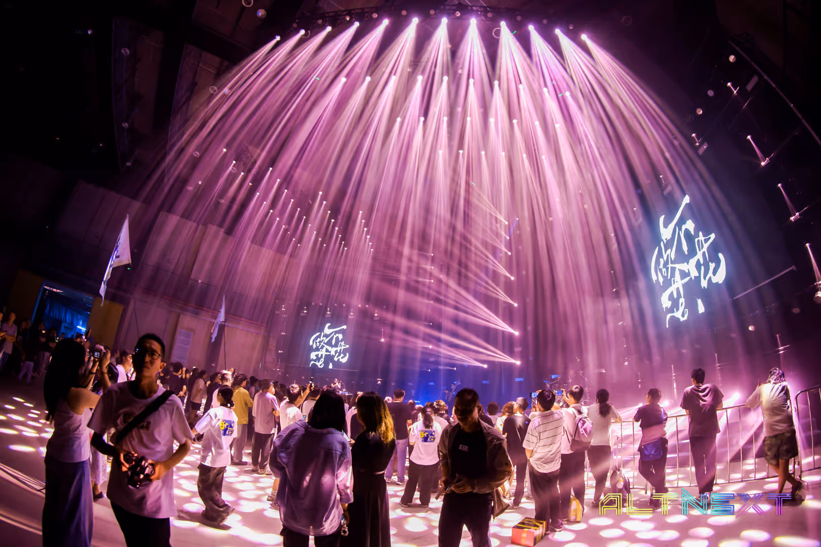 People gathered at a concert or event with purple stage lights and spotlights overhead, and a screen displaying calligraphic text - at AltNext Season 1 (2025).