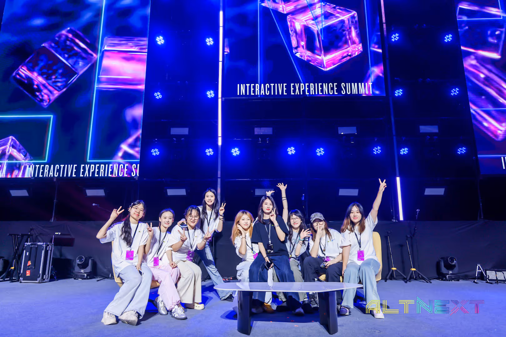 Group of nine young women sitting and posing with peace signs on a stage with glowing purple cubes displayed behind and 'Interactive Experience Summit' text visible.