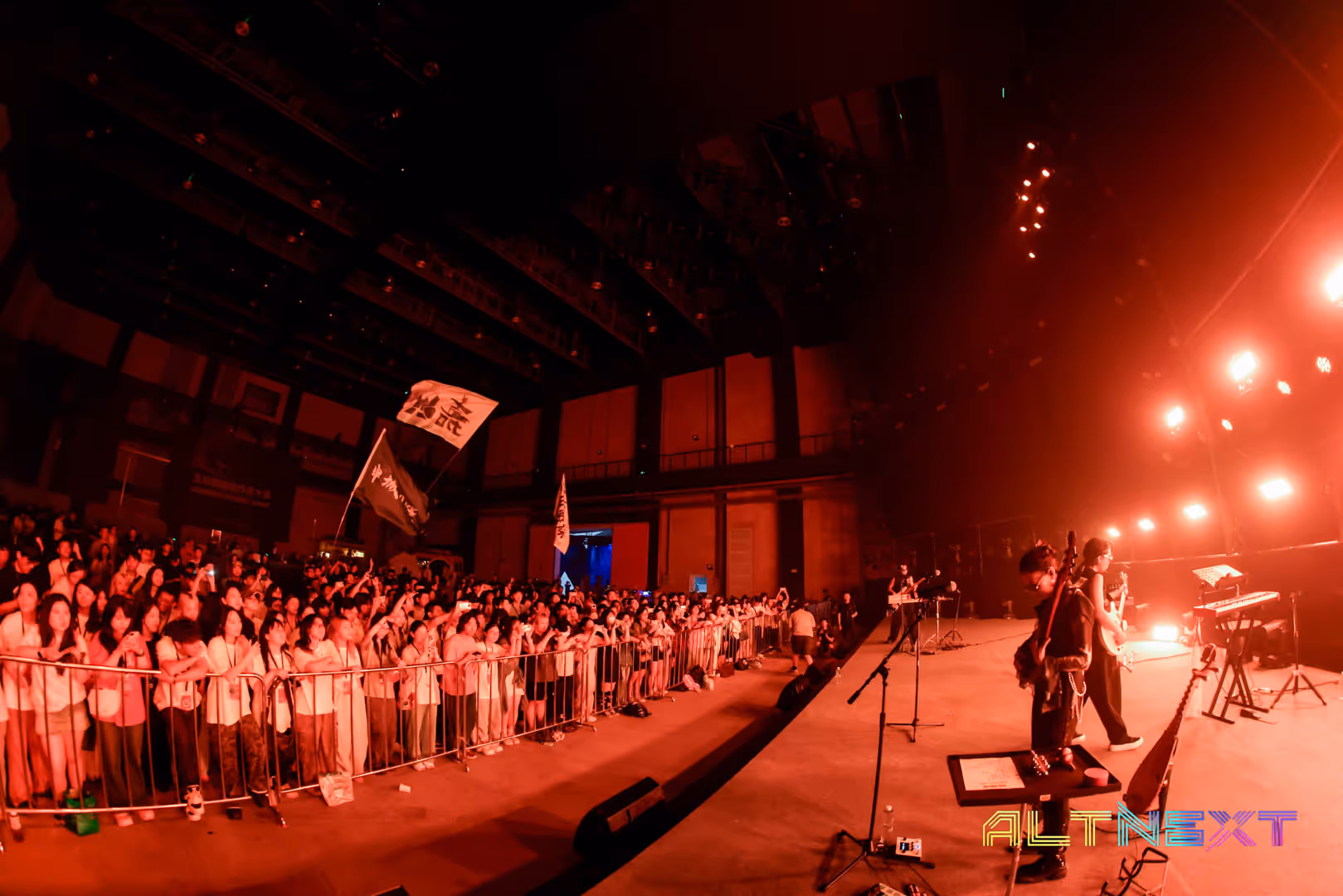 Live concert scene with a band playing on stage and a large audience behind a barricade under red lighting. Volunteers at AltNext Season 1 in 2025.