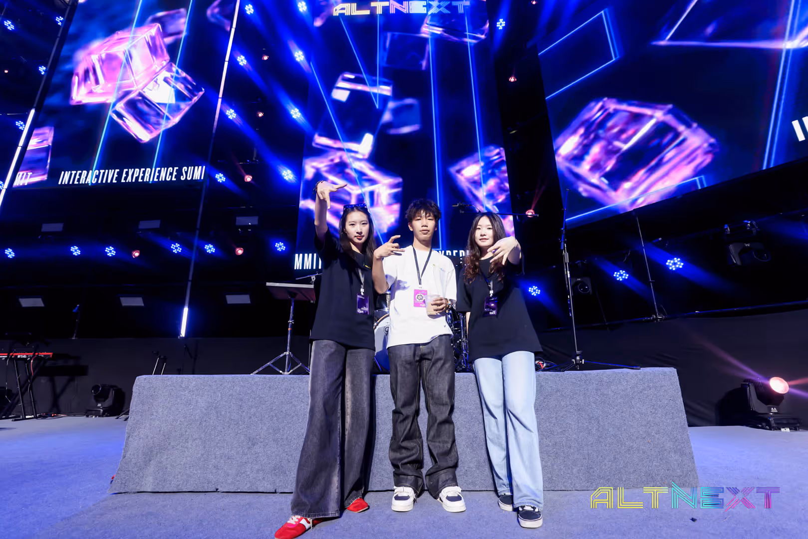 Three young people posing on stage at ALTNext event with colorful lights and digital cubes in the background.