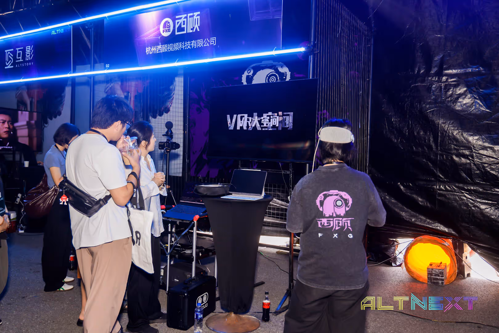 People at a tech exhibit booth with VR headset, laptops, and a screen displaying Chinese text at ALT NEXT event.