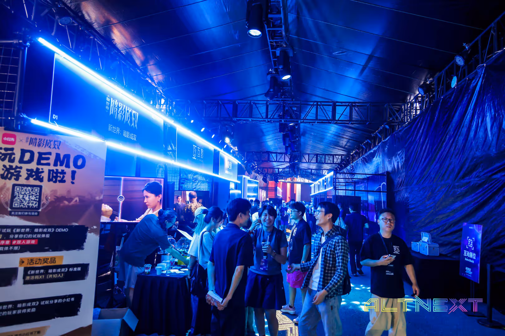 Crowd of people interacting and walking inside a neon-lit exhibition hall with a demo game booth and ALT NEXT branding.