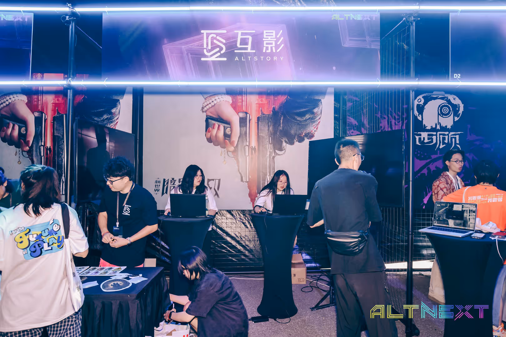 People interacting and working on laptops at the ALTSTORY booth with large banner showing hands holding guns at the ALTNEXT event.