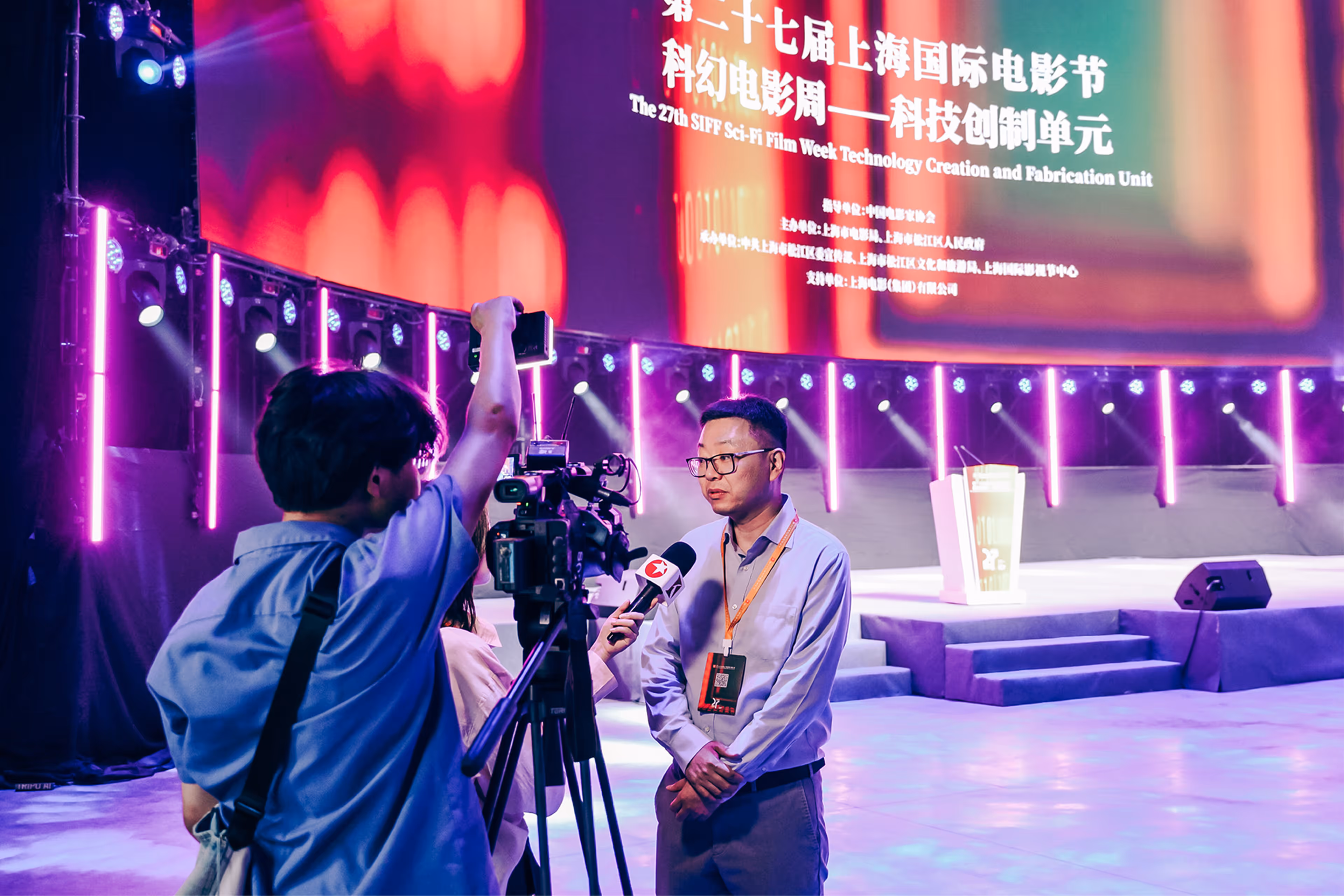 Presenter Yachen Song speaking on stage with 3D figurine models and text about the future of 3D on a large screen behind him, illuminated by pink and blue neon lights - at AltNext Edition 1 in 2025.