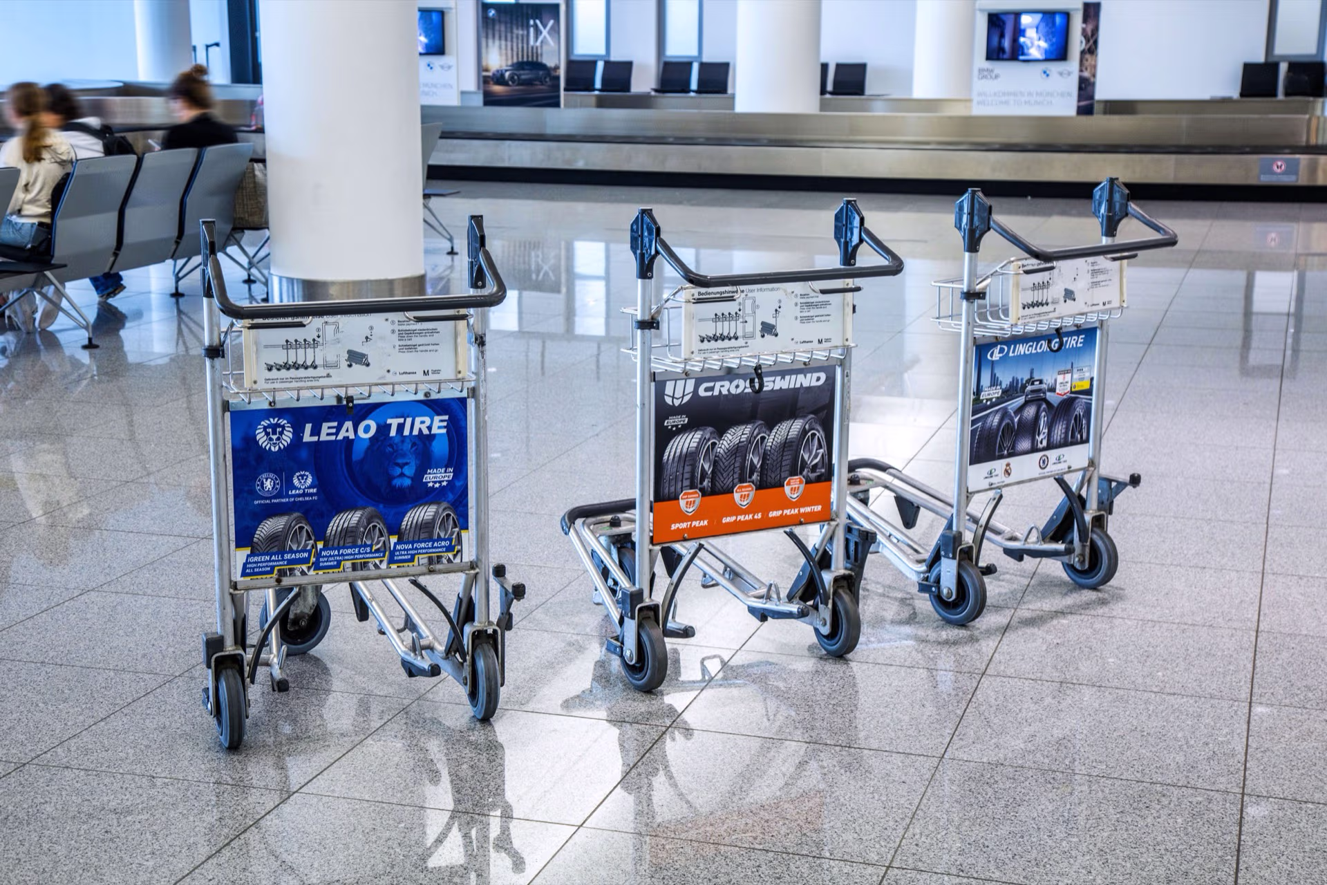 Drei Gepäckwagen im Flughafen München mit Werbeanzeigen für Leao Tire, Crosswind und Linglong Tire Reifen.